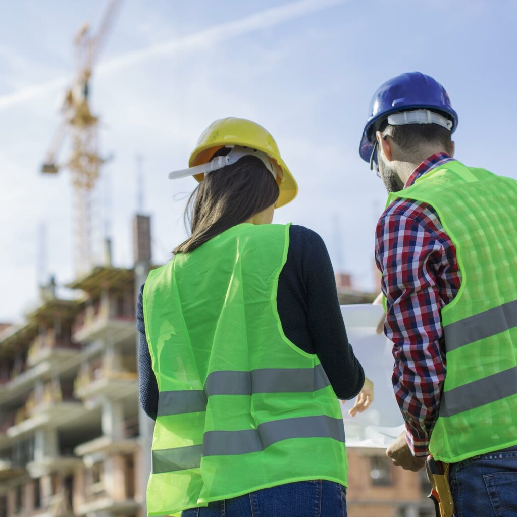 Photo of two people wearing safety gear, checking printed plans. There is a construction site and crane in the background.